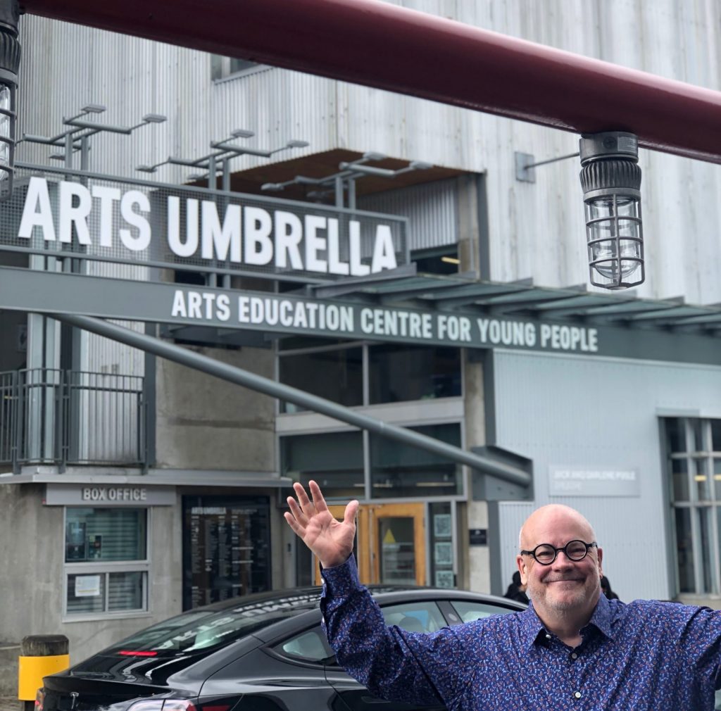 Man standing with arms up in excitement by a sign that says 'Arts Umbrella'
